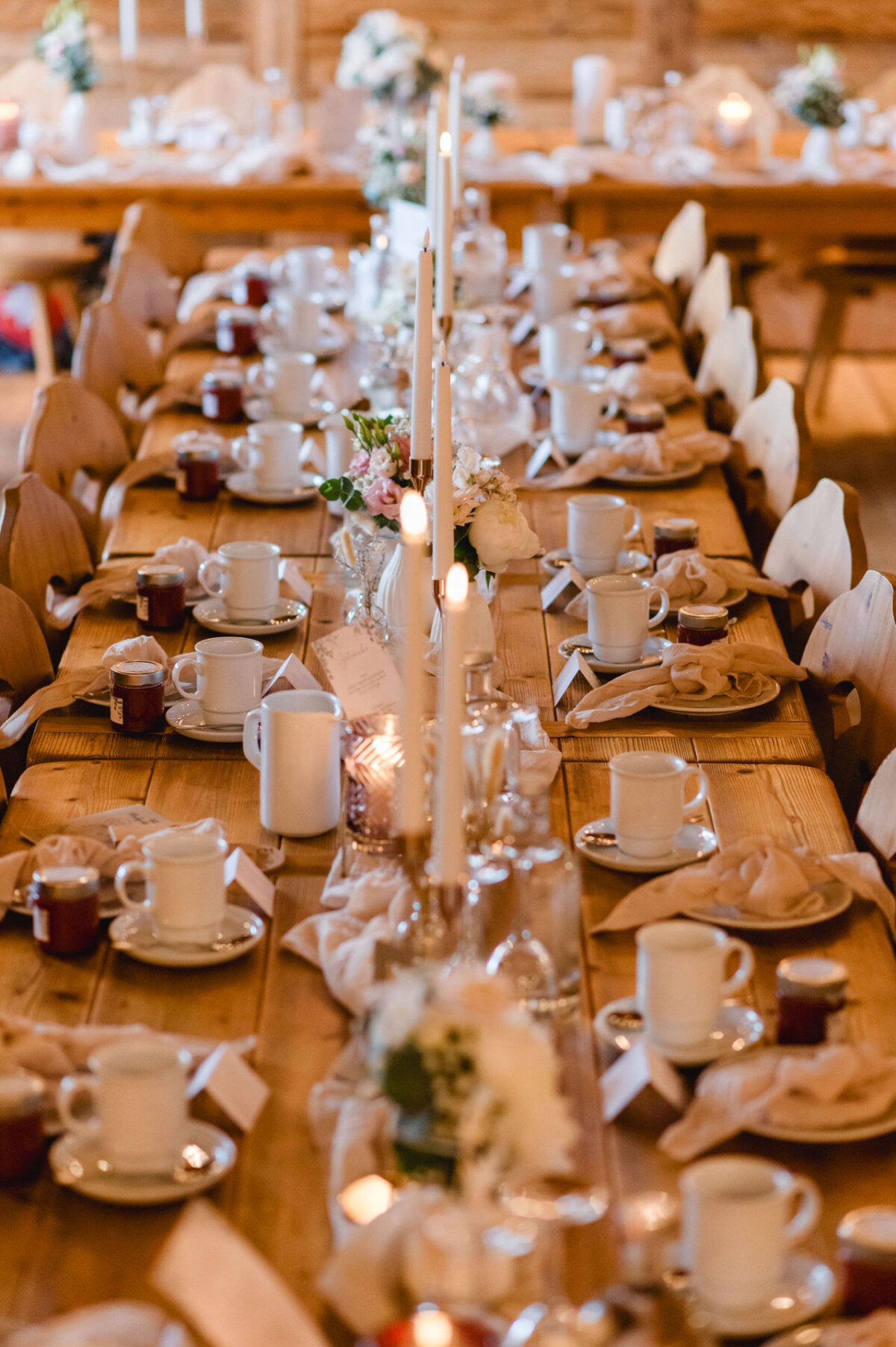 christian-lorenz.net Ein langer Holztisch ist für ein Abendessen mit weißen Tassen, Tellern, Gläsern, Kerzen und Blumendekorationen gedeckt. Neben den Tellern liegen gefaltete Servietten.