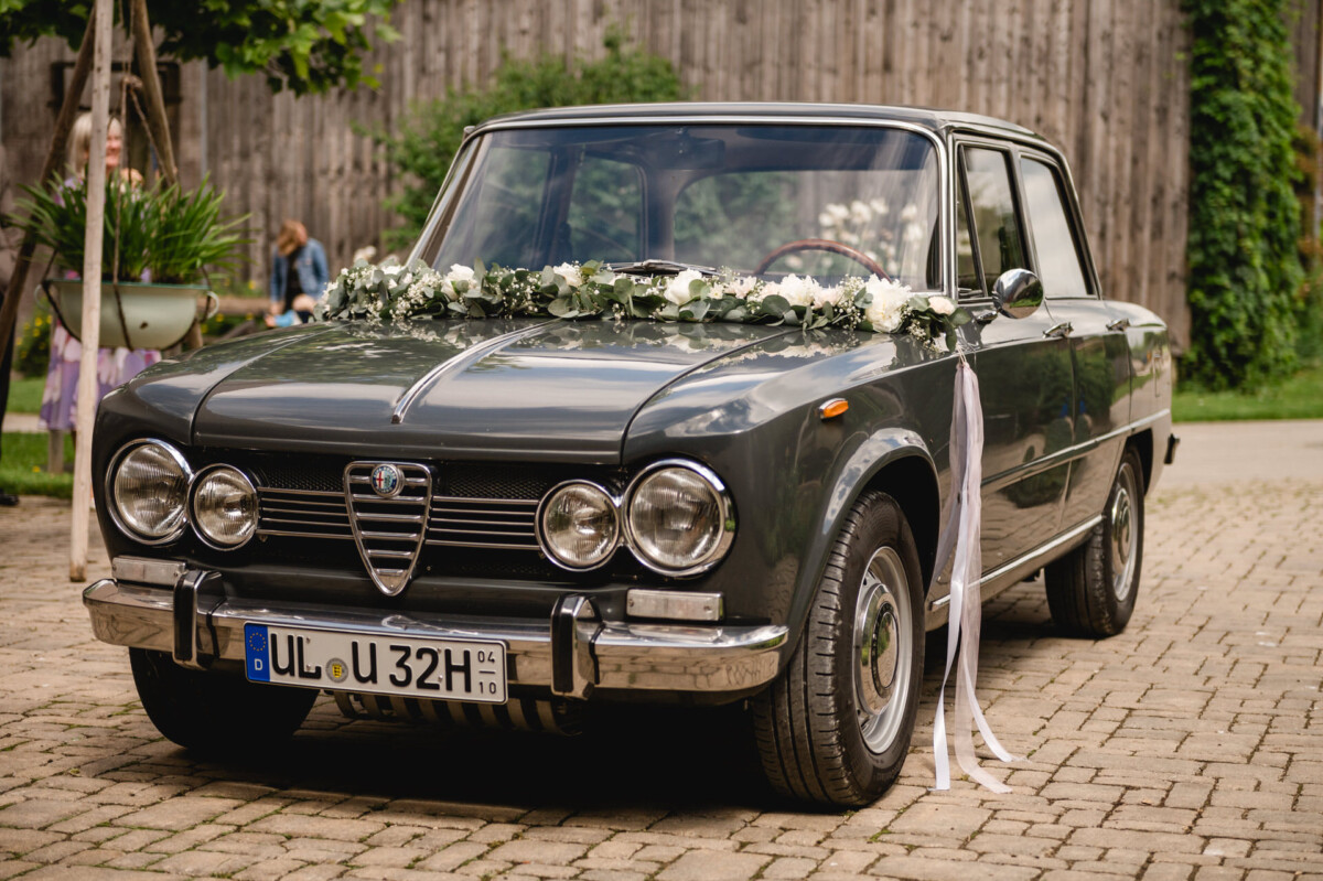 Auf einem Kopfsteinpflaster parkt ein dunkelgrünes Oldtimer, das mit weißen Blumen und Bändern geschmückt ist.
