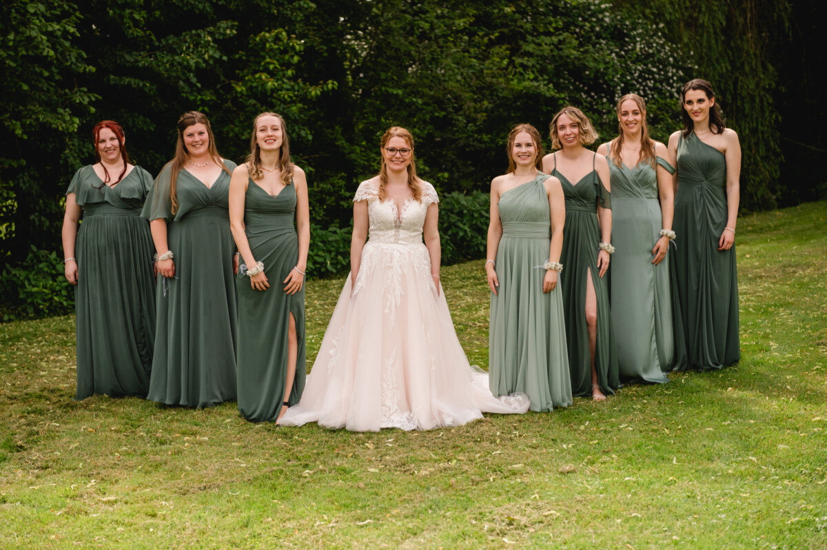 Eine Braut in einem weißen Kleid steht auf einer Wiese, umgeben von sieben Brautjungfern in grünen Kleidern. Im Hintergrund sind Bäume zu sehen.