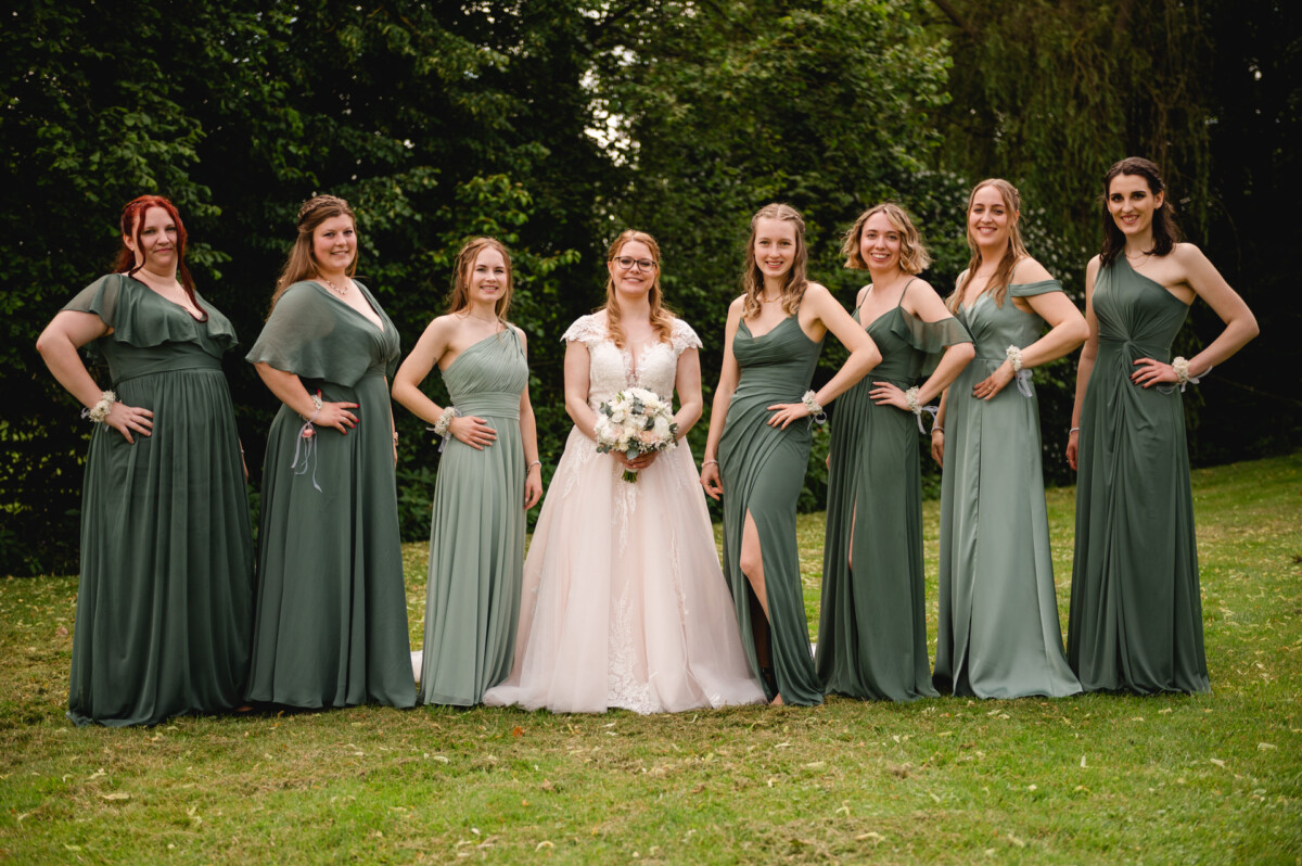 Eine Braut in einem weißen Kleid steht mit sieben Brautjungfern in grünen Kleidern auf einer Rasenfläche, umgeben von Bäumen.
