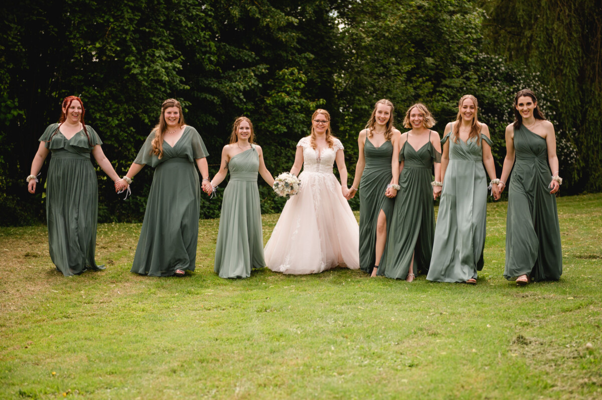 Eine Braut in einem weißen Kleid und sieben Brautjungfern in grünen Kleidern gehen Hand in Hand über das Gras, umgeben von Bäumen.