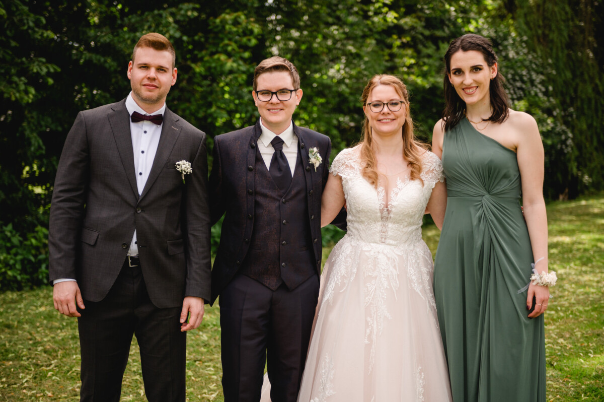 Vier Personen in formeller Kleidung stehen im Freien; zwei tragen Anzüge und zwei Kleider. Sie lächeln und posieren vor einer grünen Wiese.