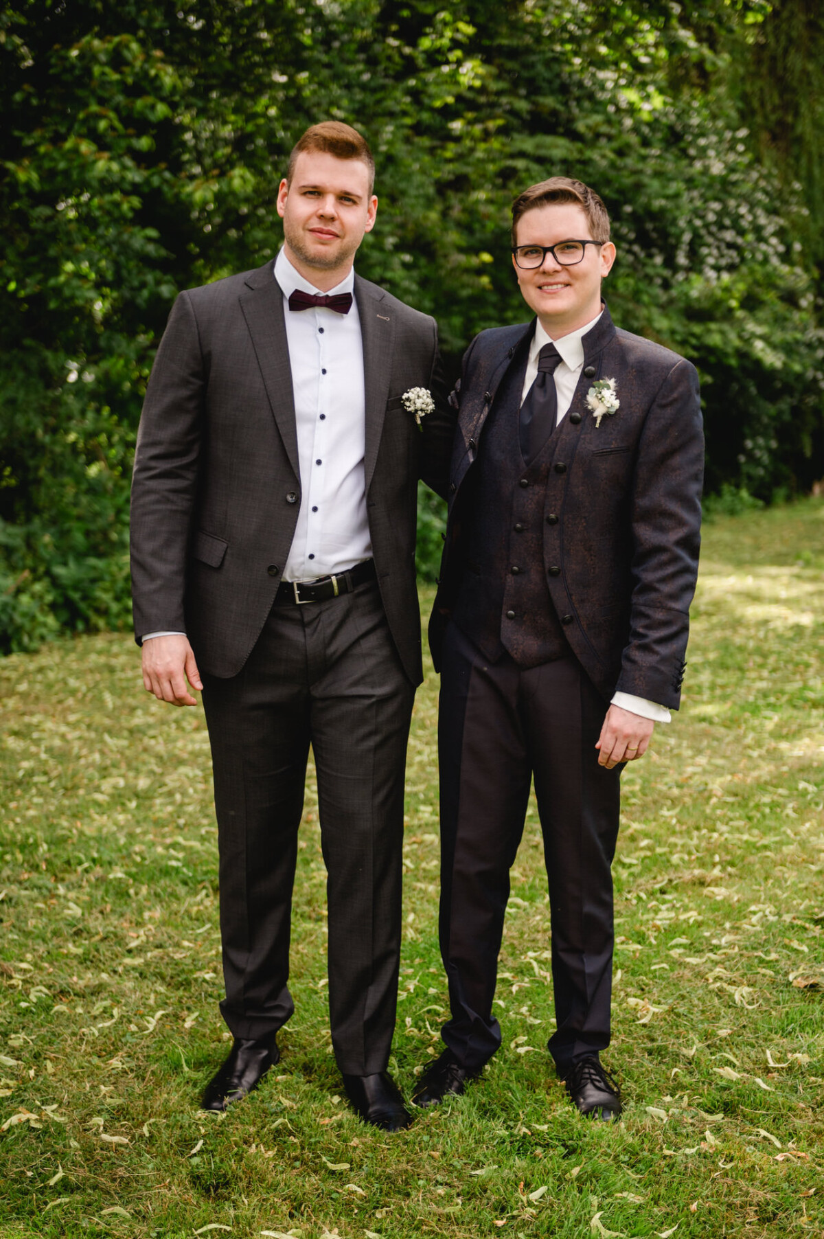 Zwei Männer in Anzügen stehen auf dem üppigen Gras vor den malerischen Bäumen des Hochzeits-Schammenhofs in Langenau bei Ulm. Mit einem herzlichen Lächeln haben beide leuchtende Blumen an ihren Jacken befestigt und fangen damit einen Moment der Freude und des Feierns ein.