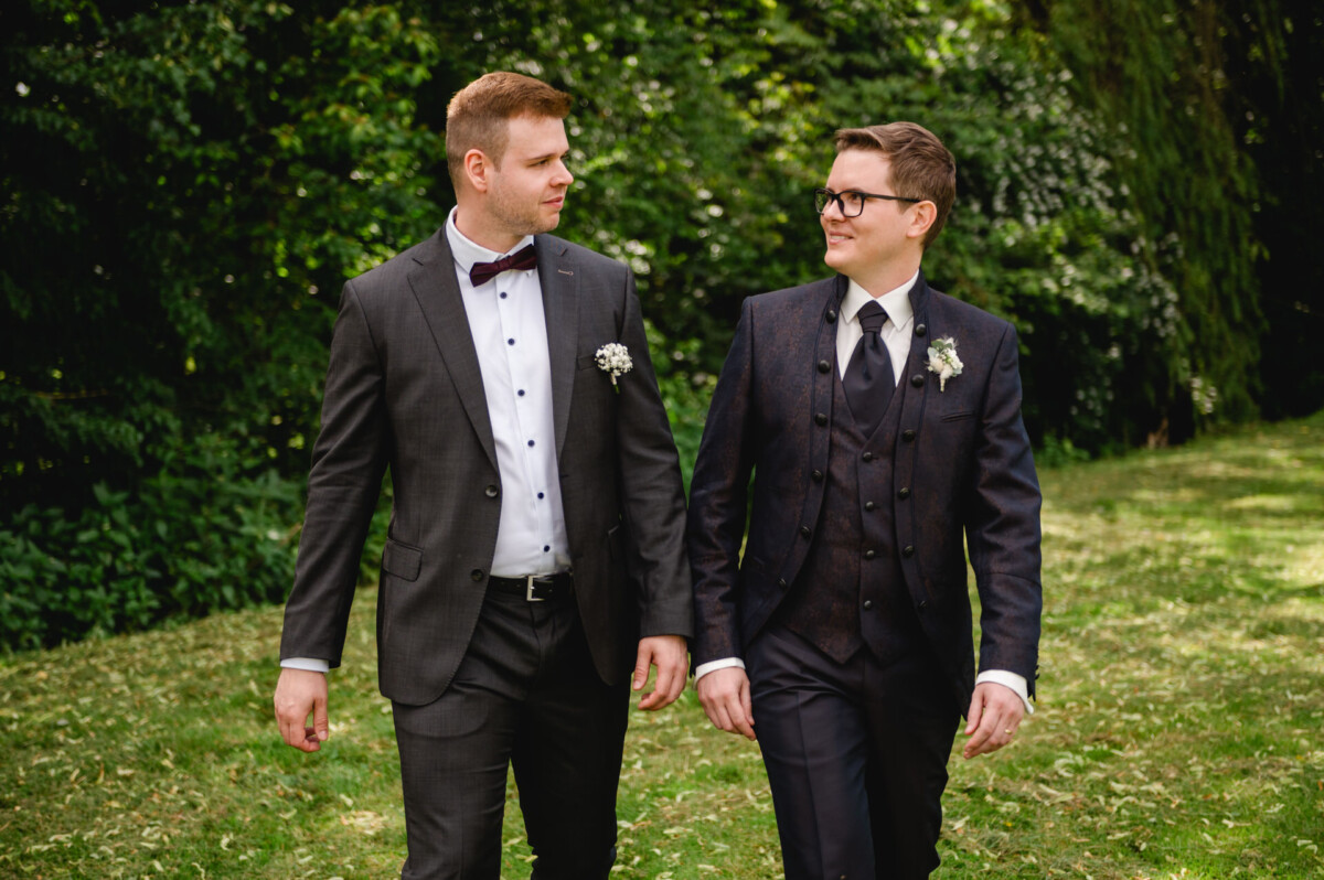 2024.10.05-Lea-Viktor-Hochzeit-Schammenhof-Langenau-bei-Ulm-00797 Zwei Männer in Anzügen gehen nebeneinander auf einem Grasweg auf dem Hochzeit Schammenhof in Langenau bei Ulm, umgeben von Bäumen. Jeder trägt eine Ansteckblume und sieht sich an.