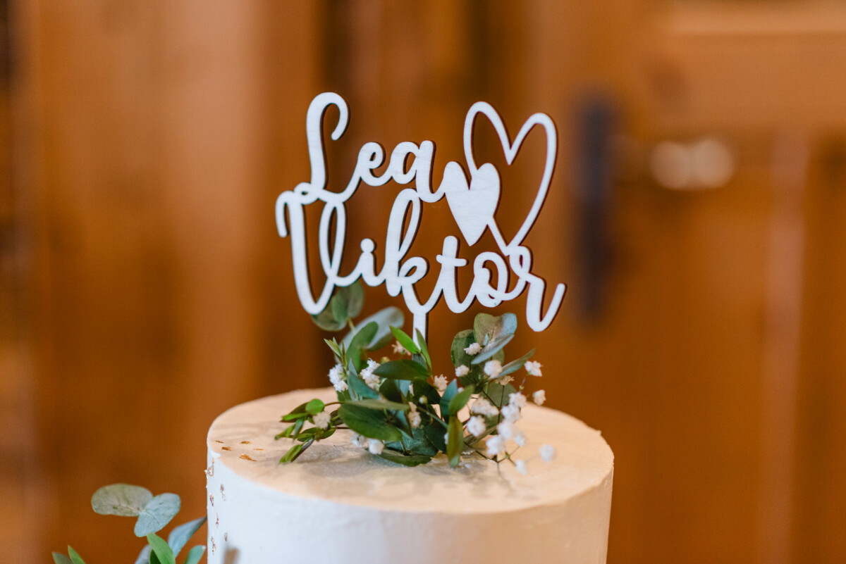 2024.10.05-Lea-Viktor-Hochzeit-Schammenhof-Langenau-bei-Ulm-00825 Eine weiße Torte, geschmückt mit einem Topper, auf dem in eleganter Schreibschrift „Lea ❤️ Viktor“ steht, umgeben von zarten Blumen und Grünpflanzen, fängt die Romantik ihrer Hochzeit auf dem Schammenhof Langenau bei Ulm perfekt ein.