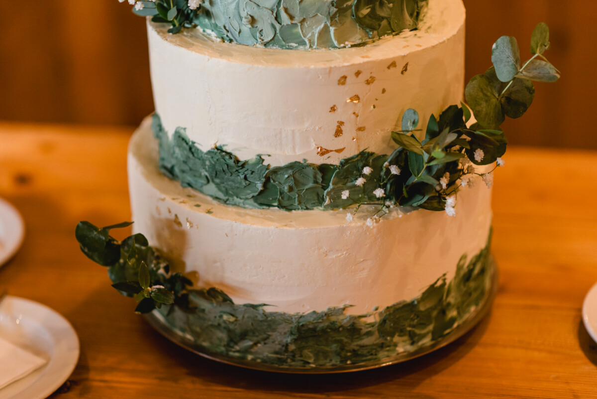 2024.10.05-Lea-Viktor-Hochzeit-Schammenhof-Langenau-bei-Ulm-00826 Eine dreistöckige weiße Torte mit grünem Strukturguss und goldenen Akzenten, geschmückt mit Blattzweigen, ziert den Holztisch. Passend für eine Hochzeit im Schammenhof Langenau bei Ulm stehen kleine Teller in der Nähe.