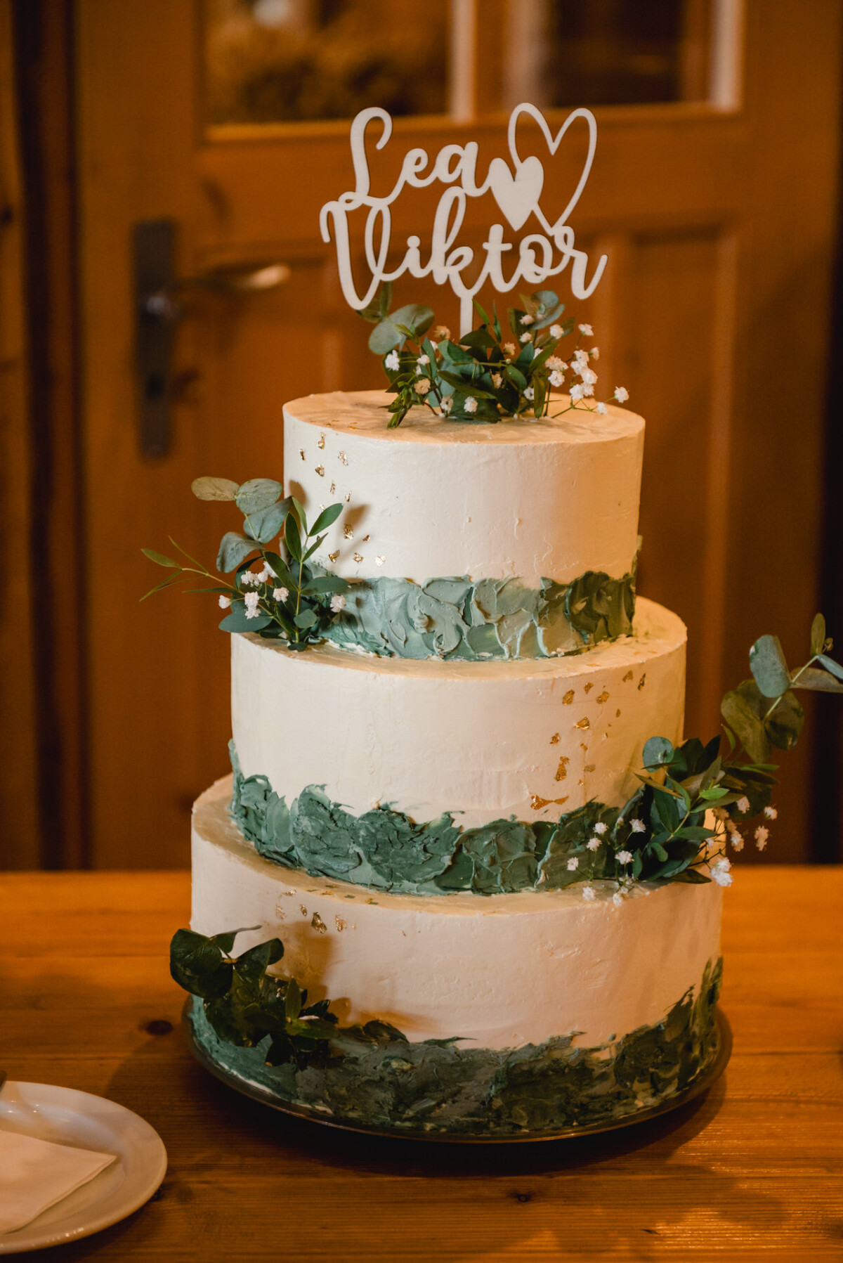 2024.10.05-Lea-Viktor-Hochzeit-Schammenhof-Langenau-bei-Ulm-00830 Eine dreistöckige Hochzeitstorte mit grünen Laubakzenten und einem Topper mit der Aufschrift „Lea ♥ Viktor“ ziert den Holztisch bei Hochzeit Schammenhof in Langenau bei Ulm.