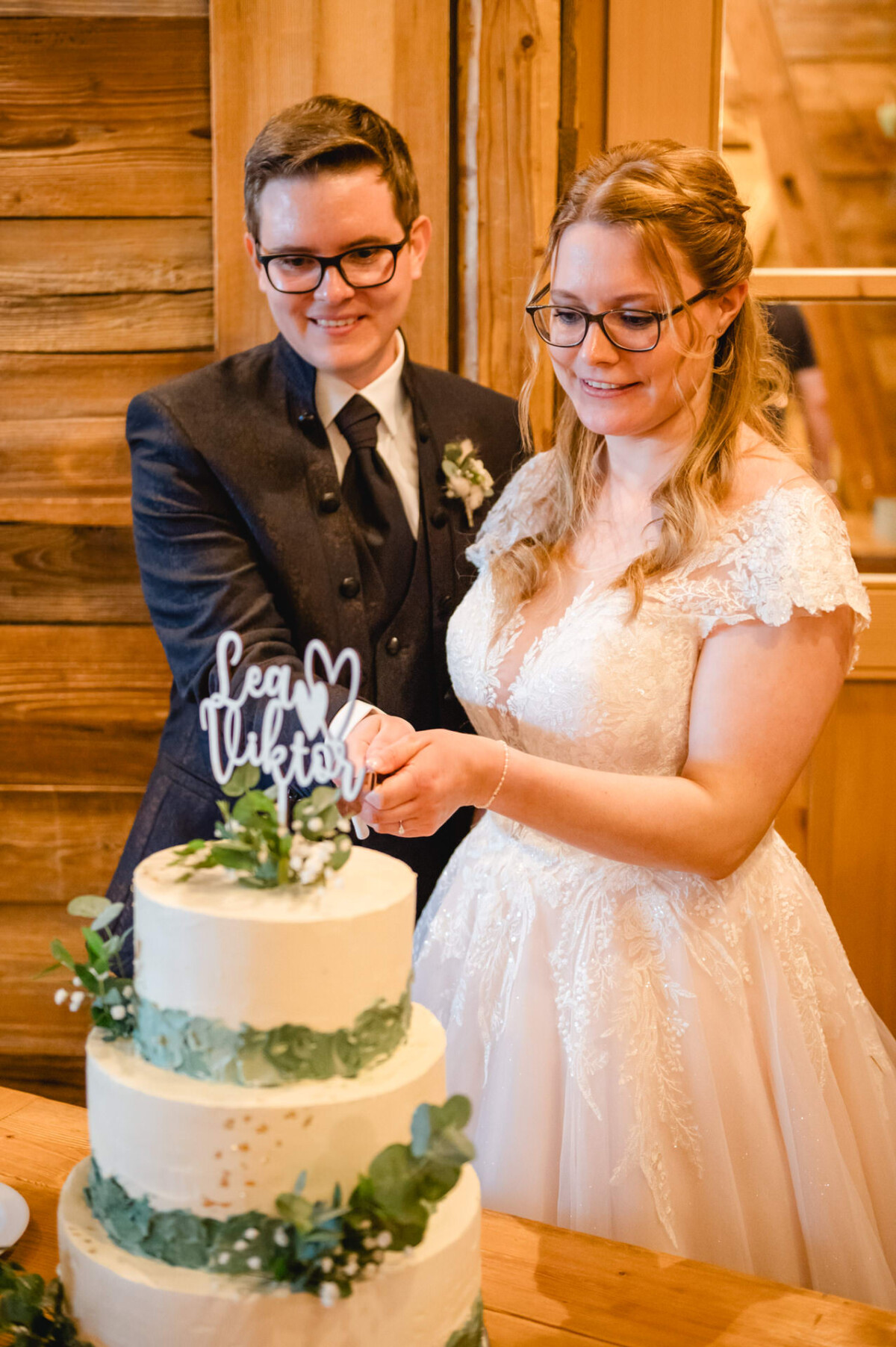 2024.10.05-Lea-Viktor-Hochzeit-Schammenhof-Langenau-bei-Ulm-00832 Bei Hochzeit Schammenhof in Langenau bei Ulm schneidet ein Paar in Abendgarderobe bei dieser Indoor-Feier elegant eine weiße, mehrstöckige Torte an, die mit Grünpflanzen geschmückt ist und auf deren Spitze ein Text zu sehen ist.