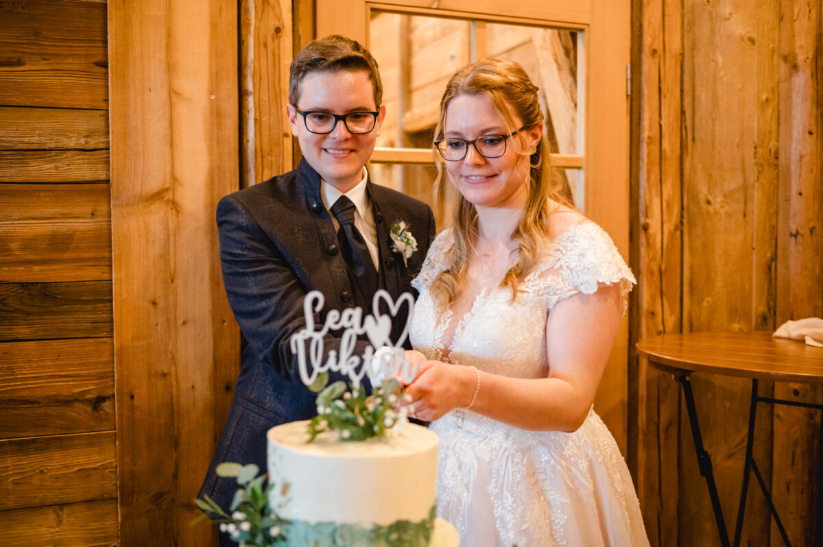 2024.10.05-Lea-Viktor-Hochzeit-Schammenhof-Langenau-bei-Ulm-00834 Ein Paar in festlicher Kleidung schneidet eine mehrstöckige Torte mit dem Aufdruck „Lena ♥ Viktor“ an, umgeben vom rustikalen Charme des Schammenhofs in Langenau bei Ulm.