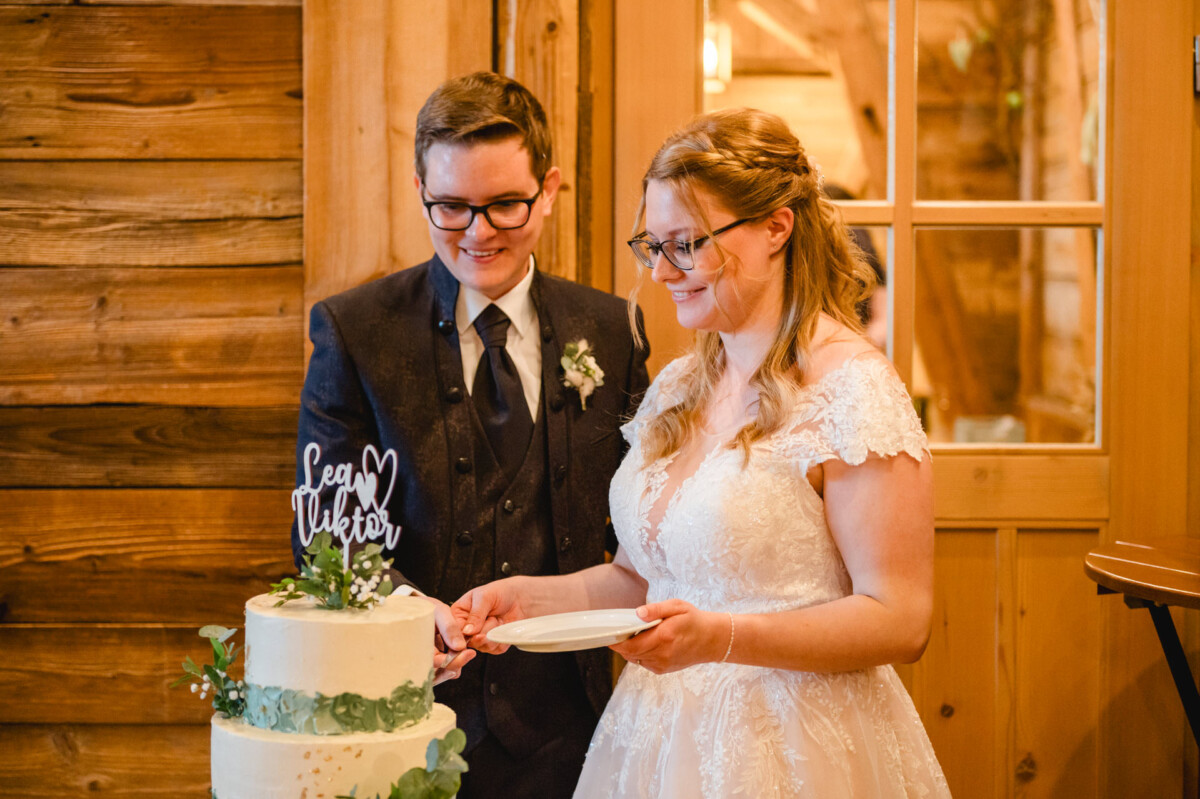 2024.10.05-Lea-Viktor-Hochzeit-Schammenhof-Langenau-bei-Ulm-00837 Braut und Bräutigam stehen neben einer mehrstöckigen Hochzeitstorte mit dem Aufsatz „Lea & Walter“, elegant gekleidet in einem rustikalen Ambiente auf dem Hochzeit Schammenhof in Langenau bei Ulm, eingerahmt von einer bezaubernden Holzkulisse.