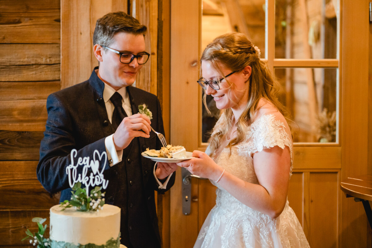 2024.10.05-Lea-Viktor-Hochzeit-Schammenhof-Langenau-bei-Ulm-00841 Auf ihrem Hochzeitsfest im Schammenhof in Langenau bei Ulm schneidet ein Paar liebevoll ein Stück Torte an und teilt sie, die mit üppigem Grün und einem Topping mit der Aufschrift „Lea“ und „Viktor“ geschmückt ist.