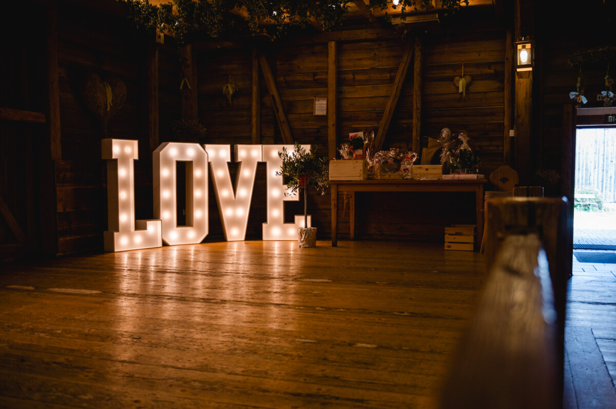 2024.10.05-Lea-Viktor-Hochzeit-Schammenhof-Langenau-bei-Ulm-00861 In einem schwach beleuchteten rustikalen Raum auf dem Hochzeit Schammenhof in der Nähe von Langenau bei Ulm ruhen große beleuchtete „LOVE“-Buchstaben auf einem Holzboden vor dem Hintergrund von Holzwänden und einem mit Blumen geschmückten Tisch.