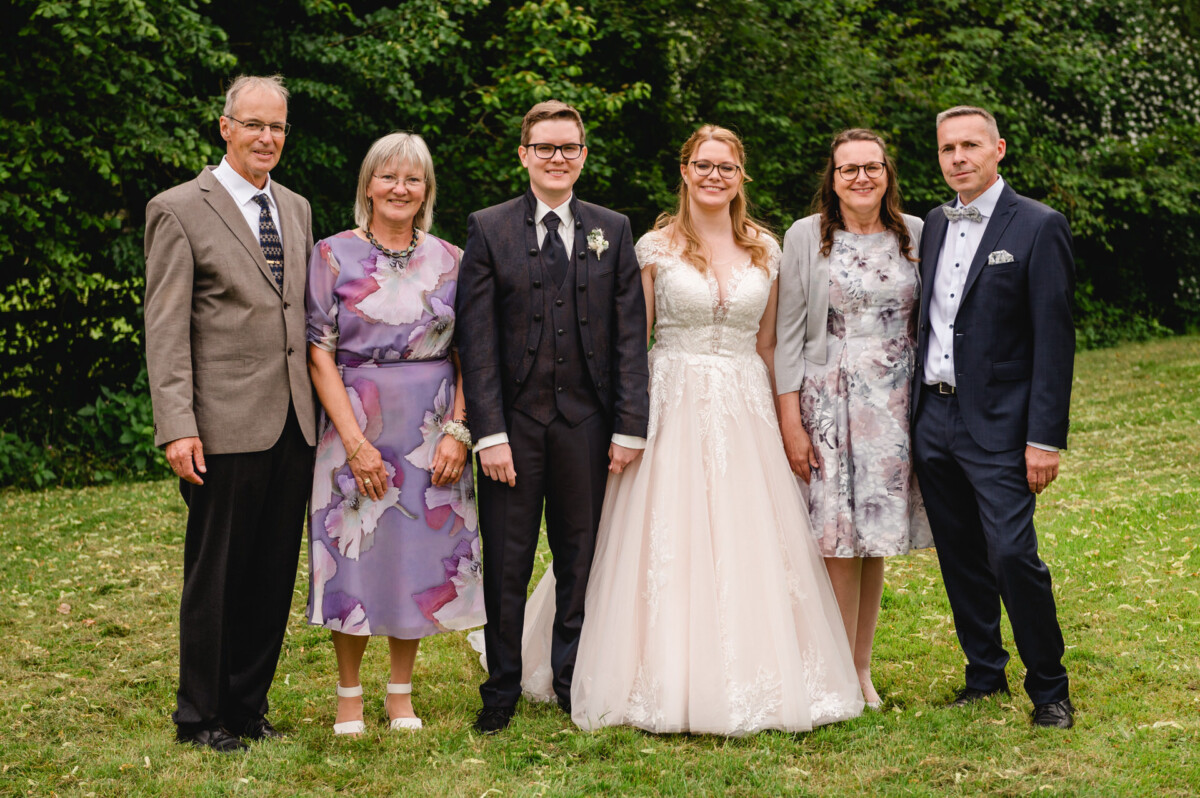 2024.10.05-Lea-Viktor-Hochzeit-Schammenhof-Langenau-bei-Ulm-00873 Ein Gruppenfoto zeigt sechs Personen auf der Wiese des Hochzeits-Schammenhofs in Langenau bei Ulm. In der Mitte ist ein Paar in Hochzeitskleidung zu sehen, umgeben von drei Frauen und zwei Männern in Abendgarderobe, alles vor einer üppigen, grünen Kulisse.