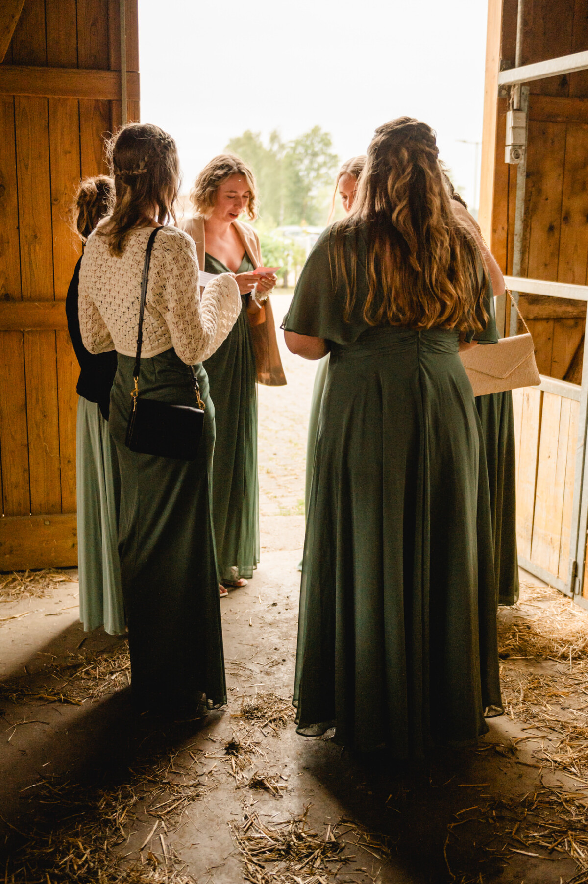 2024.10.05-Lea-Viktor-Hochzeit-Schammenhof-Langenau-bei-Ulm-00936 In der charmanten Umgebung einer Holzscheune auf dem Schammenhof in Langenau bei Ulm versammeln sich fünf Frauen in Abendgarderobe im Türrahmen und unterhalten sich intensiv.