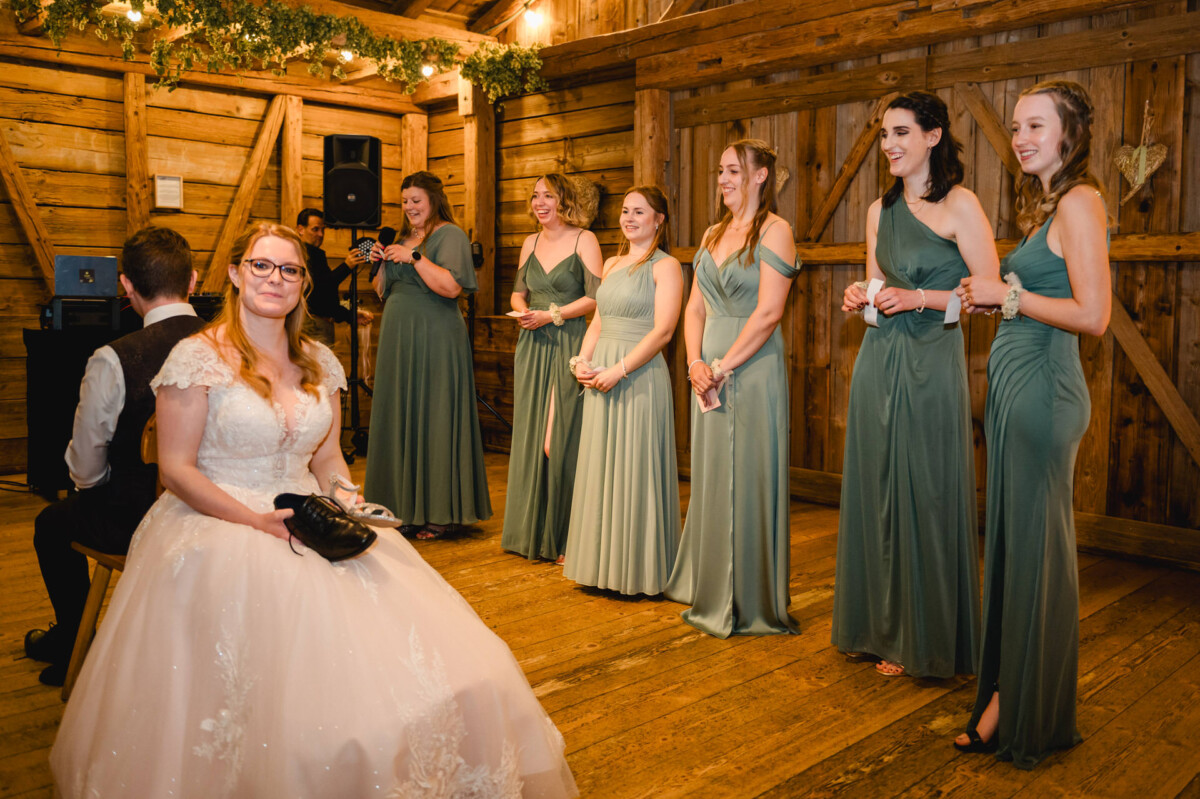 2024.10.05-Lea-Viktor-Hochzeit-Schammenhof-Langenau-bei-Ulm-00966 Bei der Hochzeit im Schammenhof in Langenau bei Ulm hält die Braut mit einem verschmitzten Grinsen einen Schuh in der Hand und sitzt im Innenbereich. Um sie herum stehen sechs Brautjungfern in eleganten grünen Kleidern, die alle in einer Reihe stehen und strahlend lächeln und einen Moment der Freude und Freundschaft einfangen.