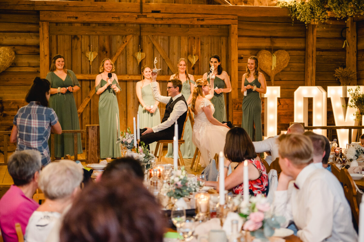 2024.10.05-Lea-Viktor-Hochzeit-Schammenhof-Langenau-bei-Ulm-00982 Bei ihrer Hochzeit auf dem Schammenhof in Langenau bei Ulm begeisterten Braut und Bräutigam die Gäste mit einem flotten Schuhspiel. Brautjungfern in grünen Kleidern stehen hinter ihnen, während ein großes, leuchtendes „LOVE“-Schild die festlichen Empfangstische in ein warmes Licht taucht.