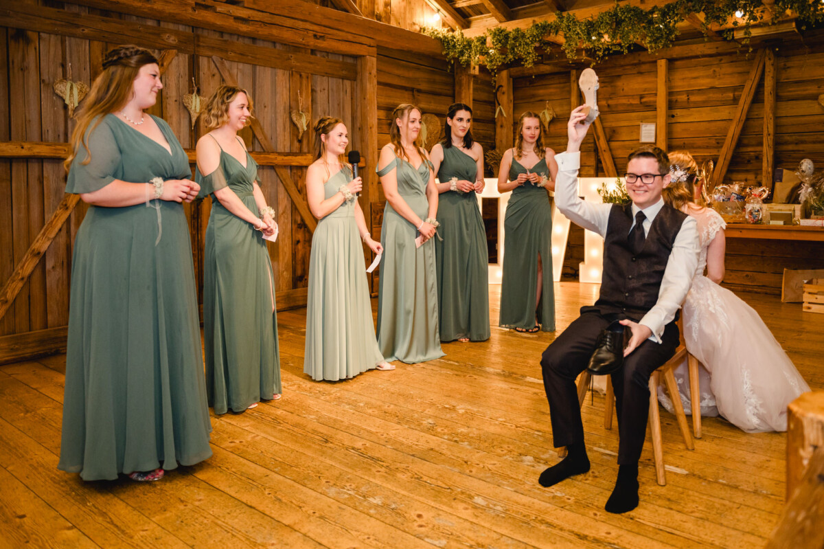 2024.10.05-Lea-Viktor-Hochzeit-Schammenhof-Langenau-bei-Ulm-00983 Bei einer Hochzeit im rustikalen Schammenhof in Langenau bei Ulm spielen ein Mann und eine Frau Rücken an Rücken das Schuhspiel. Brautjungfern in grünen Kleidern schauen zu und lachen, wodurch der Saal mit Freude erfüllt wird.