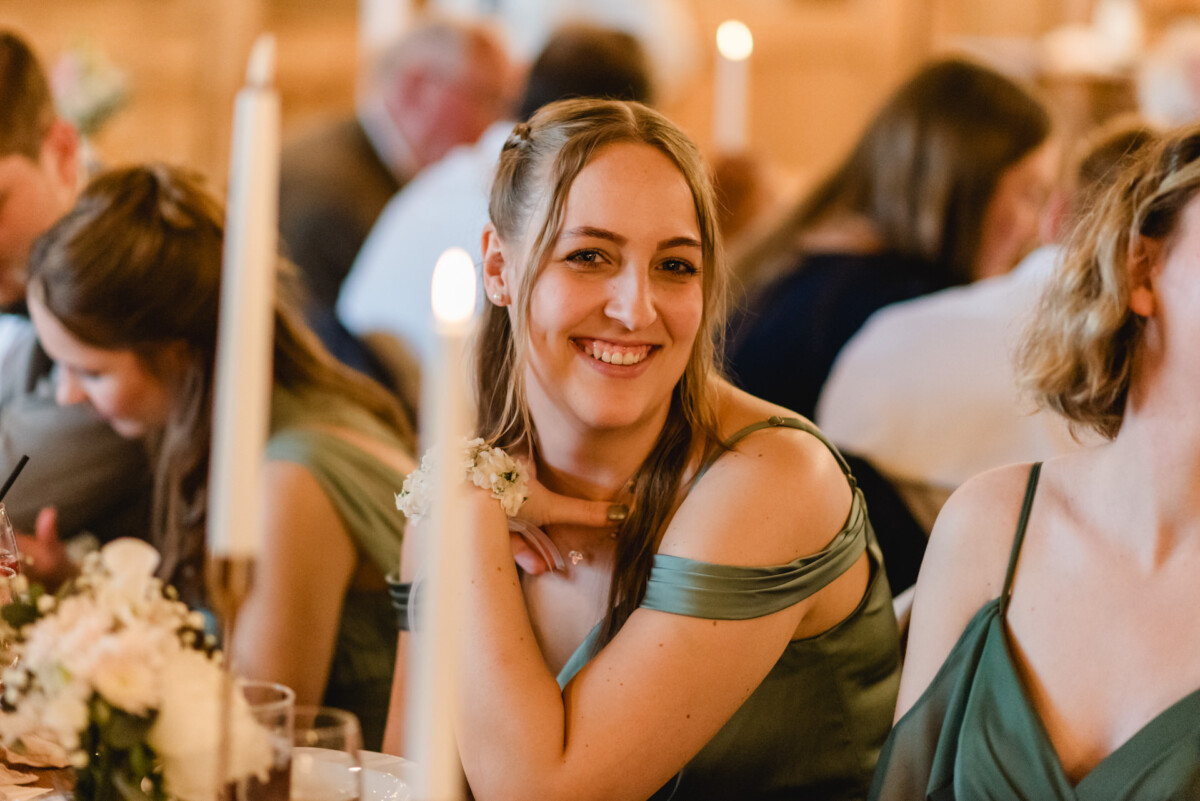 2024.10.05-Lea-Viktor-Hochzeit-Schammenhof-Langenau-bei-Ulm-01043 Bei Hochzeit Schammenhof in Langenau bei Ulm lächelt eine Frau in einem grünen Kleid, während sie an einem mit weißen Kerzen und einem Blumengesteck geschmückten Tisch sitzt, während sich im Hintergrund die Gäste unterhalten.