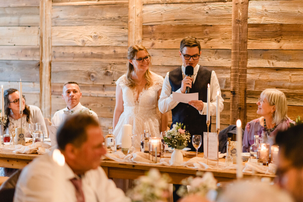2024.10.05-Lea-Viktor-Hochzeit-Schammenhof-Langenau-bei-Ulm-01088 Auf dem Hochzeits-Schammenhof in Langenau bei Ulm steht ein Paar an einem Holztisch. Der Mann hält Mikrofon und Papier in der Hand, die Frau lächelt herzlich. Gäste sitzen in der Nähe, und flackernde Kerzen schmücken die Szenerie.