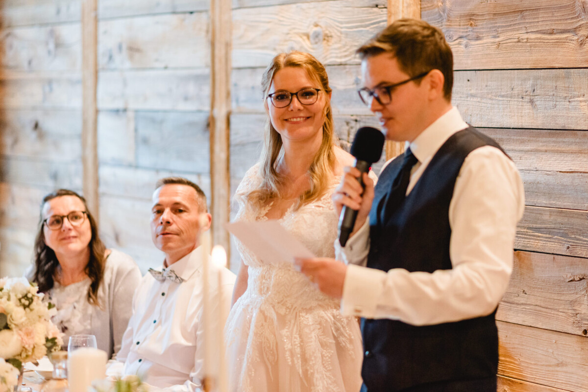 2024.10.05-Lea-Viktor-Hochzeit-Schammenhof-Langenau-bei-Ulm-01106 Ein Mann in Weste und Krawatte spricht mit einem Blatt Papier in ein Mikrofon und fängt die fröhliche Atmosphäre einer Hochzeit im Schammenhof in Langenau bei Ulm ein. Ein sitzendes Paar und eine stehende Frau im Brautkleid schauen zu, alles vor einer Holzwand im Hintergrund.