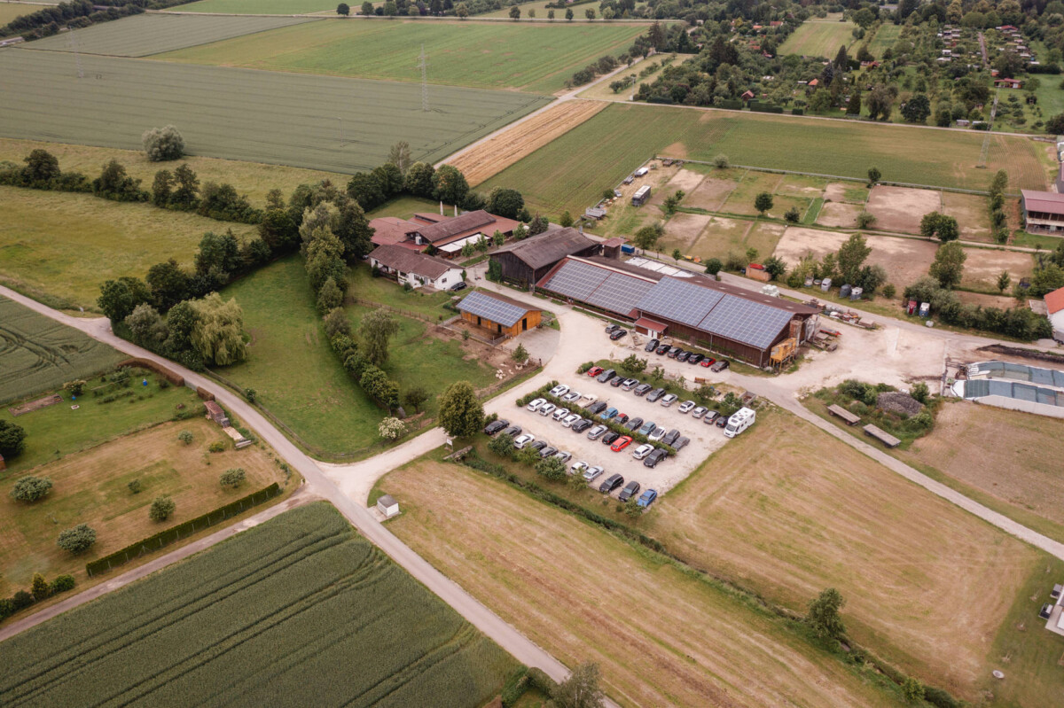 2024.10.05-Lea-Viktor-Hochzeit-Schammenhof-Langenau-bei-Ulm-01114 Eine Luftaufnahme des Hochzeit Schammenhofs in Langenau bei Ulm zeigt einen charmanten ländlichen Bauernhof mit mehreren Gebäuden und einem belebten Parkplatz, alles umgeben von üppigen grünen Feldern und Bäumen.
