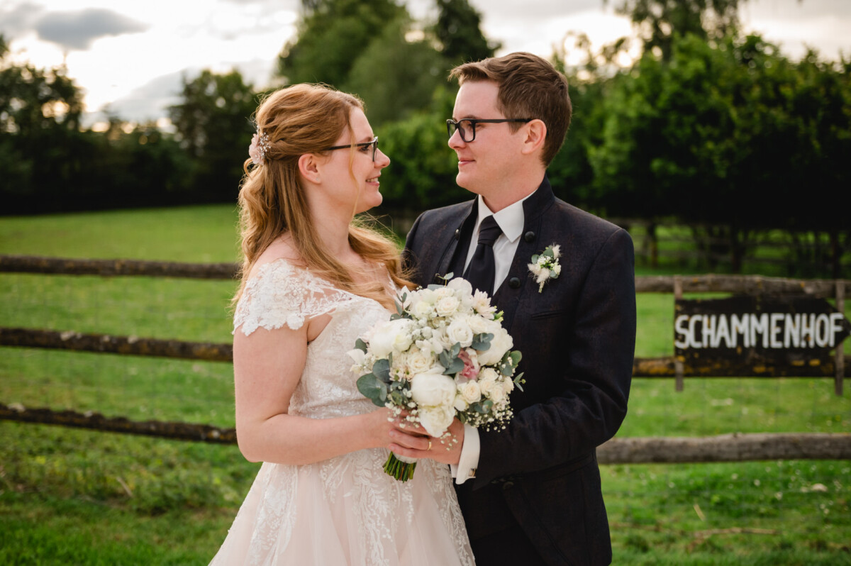 2024.10.05-Lea-Viktor-Hochzeit-Schammenhof-Langenau-bei-Ulm-01145 Auf einer malerischen Wiese auf dem Hochzeit Schammenhof in Langenau bei Ulm stehen Braut und Bräutigam zusammen und halten einen weißen Blumenstrauß in den Händen. Sie blicken sich an und lächeln herzlich. Ein Holzzaun und Bäume bilden eine bezaubernde Kulisse für diese idyllische Szene.