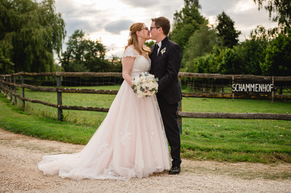 2024.10.05-Lea-Viktor-Hochzeit-Schammenhof-Langenau-bei-Ulm-01153 Braut und Bräutigam küssen sich vor dem rustikalen Holzzaun des „Schammenhof“-Schildes, umgeben von üppigem Grün bei ihrer Hochzeit in Langenau bei Ulm. Die Braut hält in ihrem weißen Kleid einen Blumenstrauß, während der Bräutigam im Anzug unter einem wolkenverhangenen Himmel neben ihr steht.