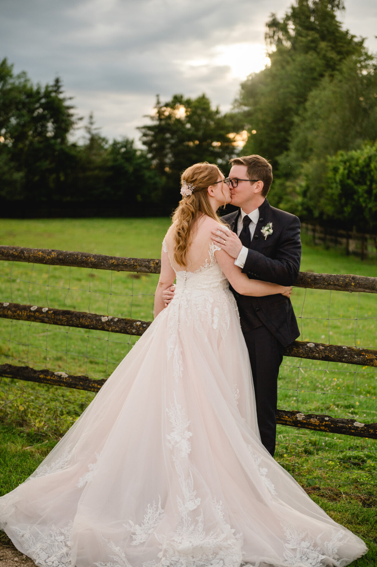 2024.10.05-Lea-Viktor-Hochzeit-Schammenhof-Langenau-bei-Ulm-01173 Ein Paar in Hochzeitskleidung küsst sich auf dem Hochzeit Schammenhof in Langenau, mit einem Holzzaun und üppigem Grün unter einem bewölkten Himmel.