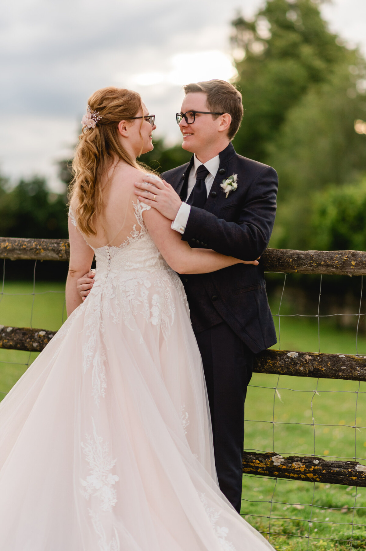 2024.10.05-Lea-Viktor-Hochzeit-Schammenhof-Langenau-bei-Ulm-01176 Auf dem malerischen Hochzeit Schammenhof bei Langenau bei Ulm stehen eine Braut im weißen Spitzenkleid und ein Bräutigam im dunklen Anzug liebevoll an einem Holzzaun inmitten üppigen Grüns.