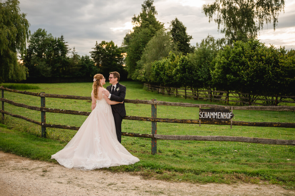 2024.10.05-Lea-Viktor-Hochzeit-Schammenhof-Langenau-bei-Ulm-01177 Ein Paar in festlicher Kleidung steht an einem Holzzaun auf einer Wiese beim Hochzeitshof Schammenhof. Auf dem Schild steht „Schammenhof“, vor der Kulisse der Bäume von Langenau bei Ulm und einem bewölkten Himmel.