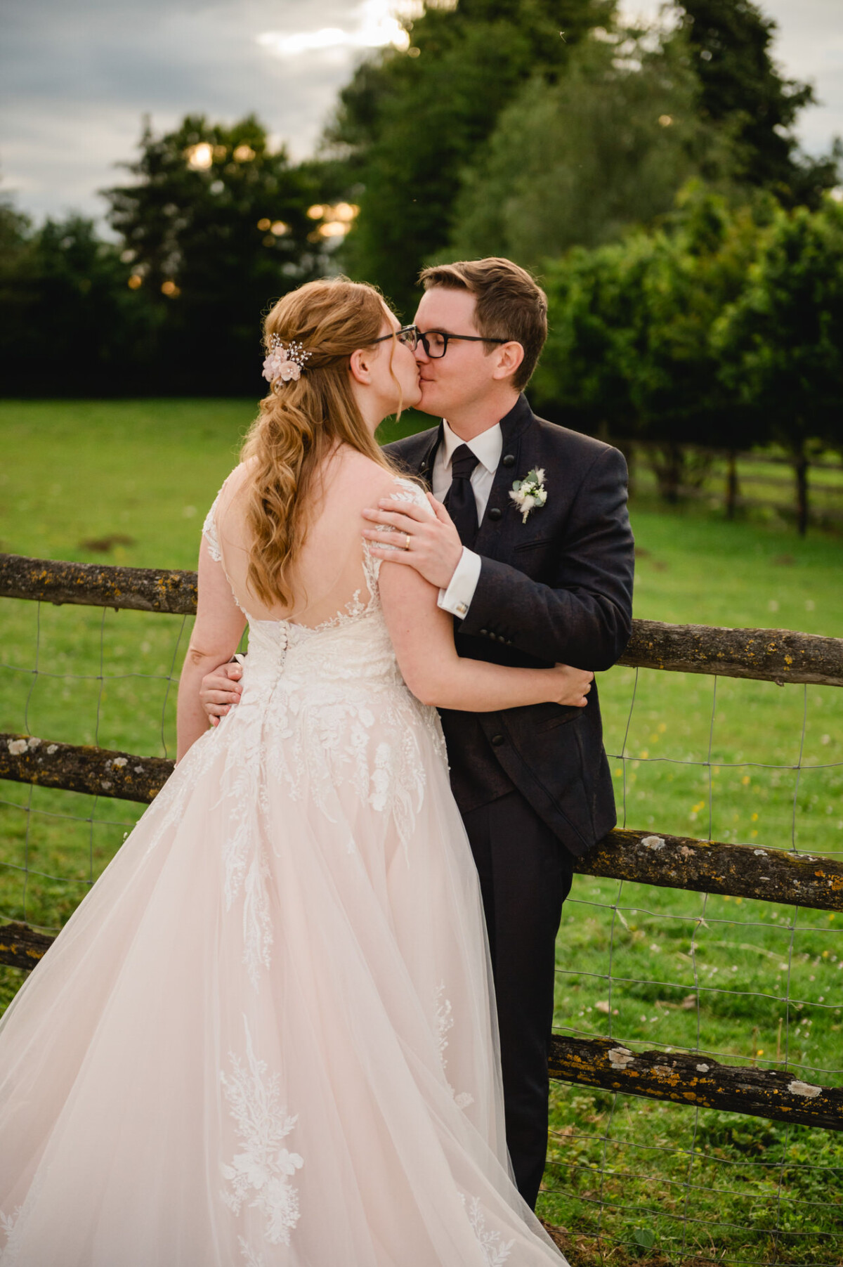 2024.10.05-Lea-Viktor-Hochzeit-Schammenhof-Langenau-bei-Ulm-01180 Braut und Bräutigam küssen sich vor einem Holzzaun, dahinter Wiesen und Bäume auf dem Hochzeit Schammenhof in Langenau bei Ulm. Die Braut glänzt in ihrem Spitzenbrautkleid, der Bräutigam sieht in seinem Anzug elegant aus.