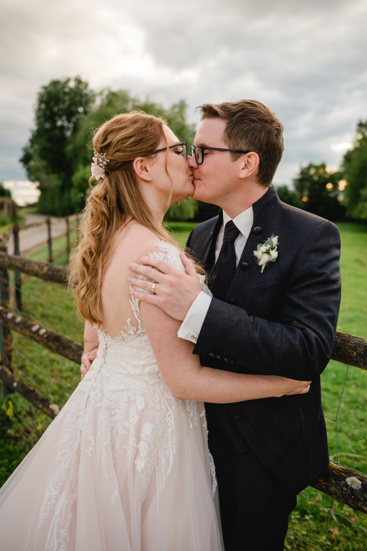2024.10.05-Lea-Viktor-Hochzeit-Schammenhof-Langenau-bei-Ulm-01184 Braut und Bräutigam küssen sich draußen an einem Zaun, umgeben vom üppigen Grün des Hochzeits-Schammenhofs in Langenau bei Ulm. Die Braut sieht in ihrem weißen Spitzenkleid umwerfend aus, während der Bräutigam in seinem dunklen Anzug elegant aussieht.