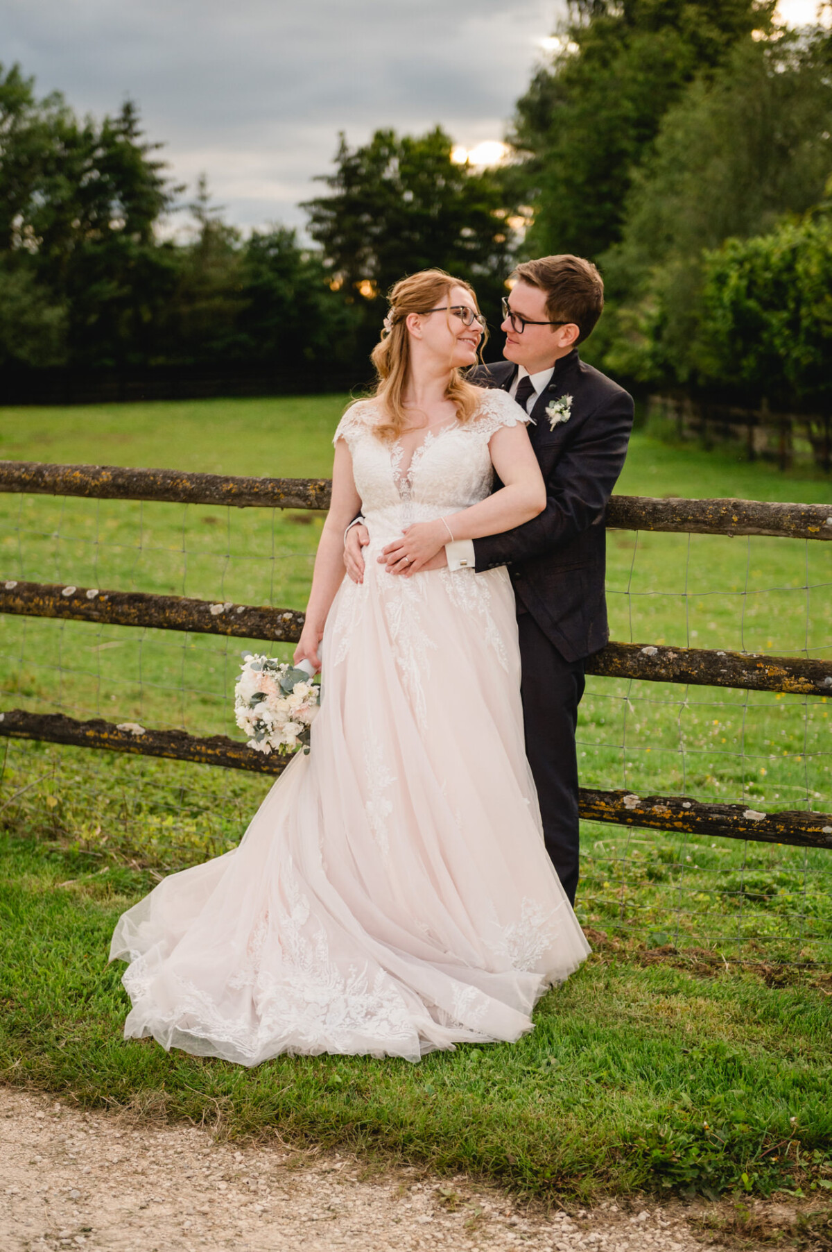 2024.10.05-Lea-Viktor-Hochzeit-Schammenhof-Langenau-bei-Ulm-01192 Braut und Bräutigam stehen gemeinsam an einem Holzzaun auf einer Wiese auf dem Hochzeitsplatz Schammenhof in Langenau bei Ulm. Die Braut hält einen Blumenstrauß in der Hand und trägt ein weißes Kleid, während der Bräutigam einen dunklen Anzug trägt. Im Hintergrund sind Bäume zu sehen.