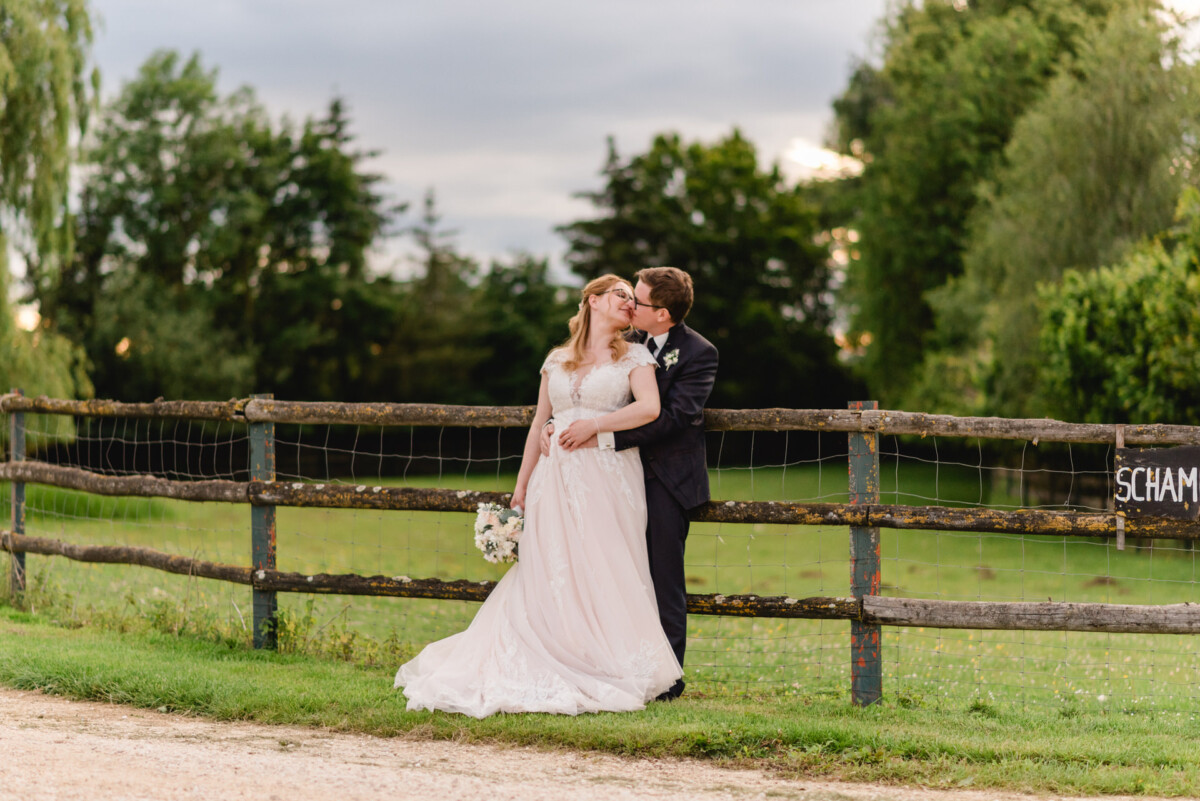 christian-lorenz.net Ein Paar in Hochzeitskleidung steht an einem Holzzaun. Der Bräutigam küsst die Braut auf die Wange, während sie einen Blumenstrauß hält. Im Hintergrund sind grüne Bäume und ein bewölkter Himmel zu sehen.