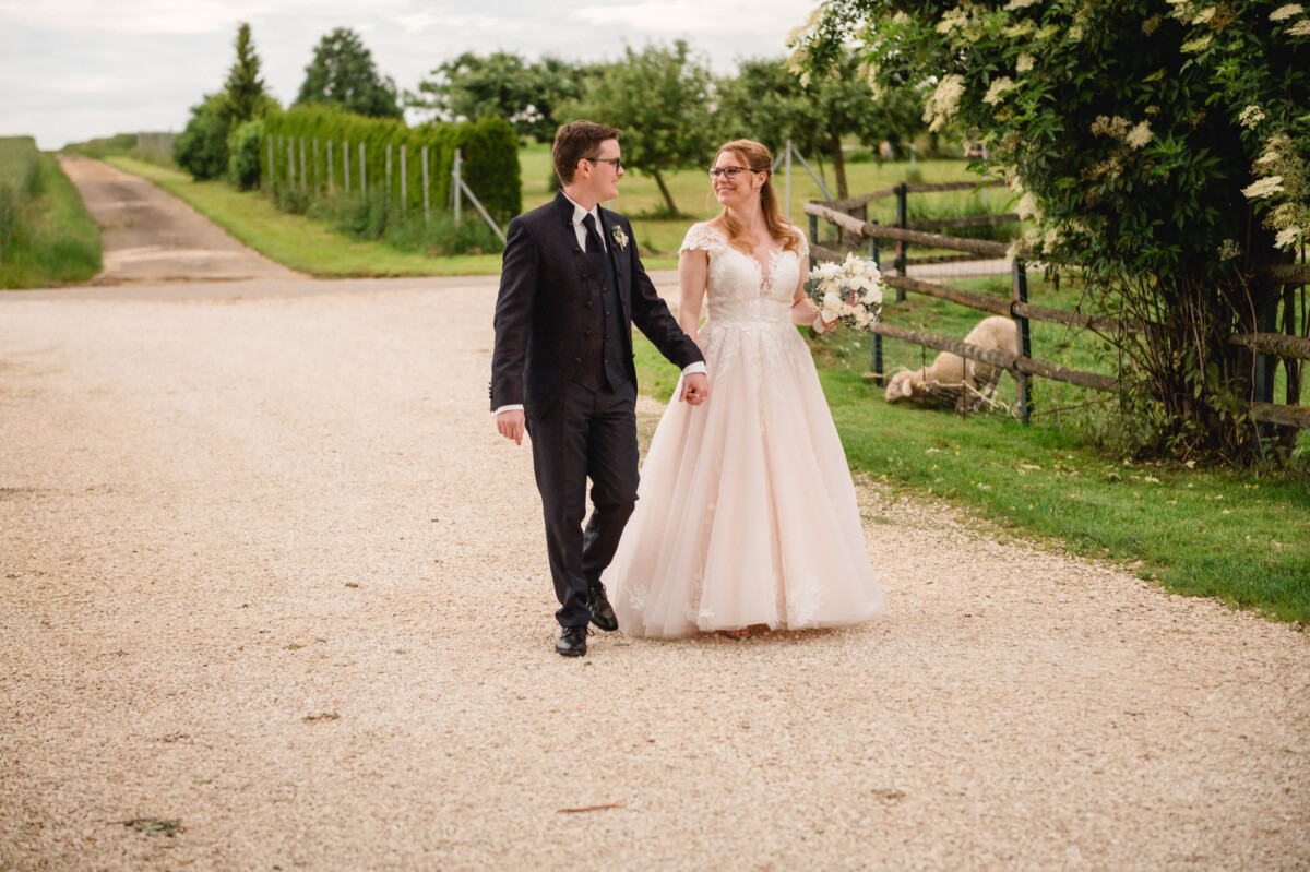 Ein Brautpaar geht Hand in Hand auf einem Schotterweg auf dem Hochzeit Schammenhof, umgeben von Grün und einem Holzzaun, während im Hintergrund sanft grasende Schafe in der Nähe von Langenau bei Ulm grasen. Ein Brautpaar geht Hand in Hand auf einem Schotterweg auf dem Hochzeit Schammenhof, umgeben von Grün und einem Holzzaun, während im Hintergrund sanft grasende Schafe in der Nähe von Langenau bei Ulm grasen.