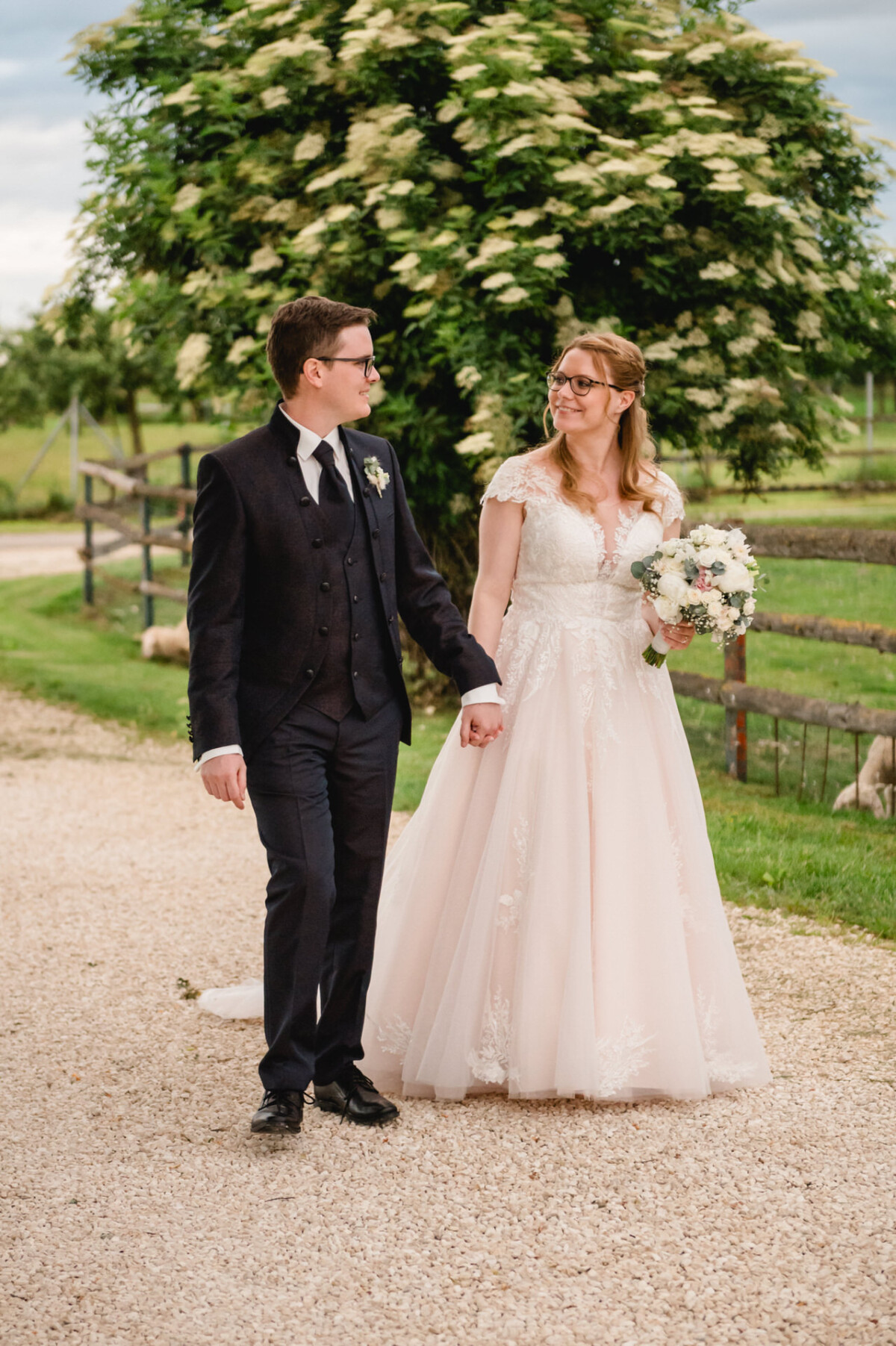 Ein Paar geht Händchen haltend in seiner Hochzeitskleidung einen Kiesweg entlang auf dem Hochzeit Schammenhof. Sie lächeln sich an, mit viel Grün und einem Holzzaun im Hintergrund, der die bezaubernde Atmosphäre von Langenau bei Ulm einfängt. Ein Paar geht Händchen haltend in seiner Hochzeitskleidung einen Kiesweg entlang auf dem Hochzeit Schammenhof. Sie lächeln sich an, mit viel Grün und einem Holzzaun im Hintergrund, der die bezaubernde Atmosphäre von Langenau bei Ulm einfängt.