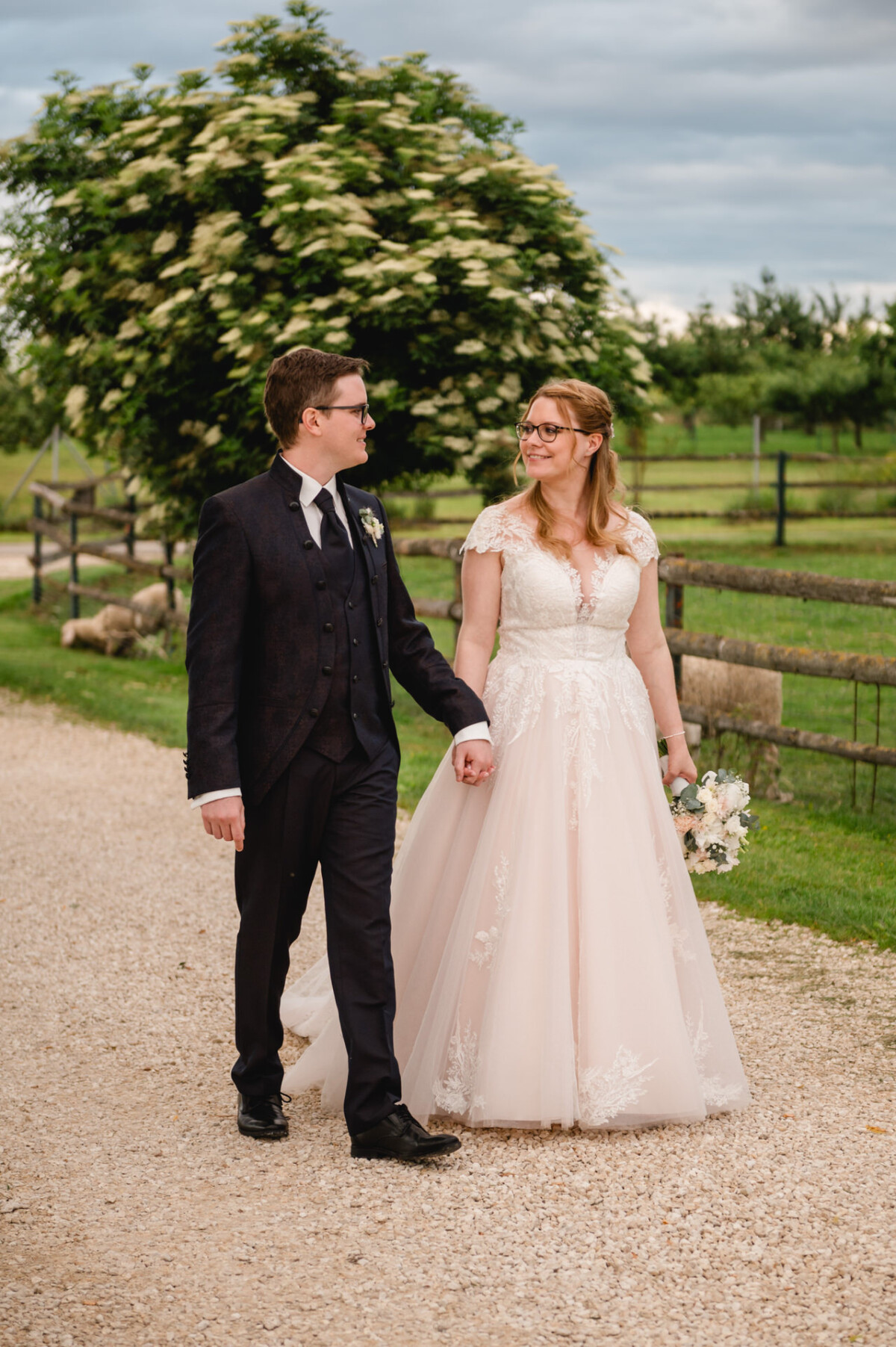 Ein Paar im Hochzeitskleid schlendert Hand in Hand auf einem Kiesweg auf dem Hochzeit Schammenhof, umgeben von Grün und Holzzäunen in der Nähe von Langenau bei Ulm. Ein Paar im Hochzeitskleid schlendert Hand in Hand auf einem Kiesweg auf dem Hochzeit Schammenhof, umgeben von Grün und Holzzäunen in der Nähe von Langenau bei Ulm.