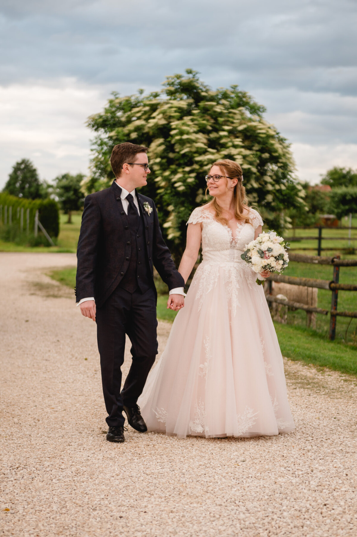 Ein Brautpaar in Abendgarderobe geht Hand in Hand auf einem Schotterweg beim Hochzeit Schammenhof, während Grün und ein bewölkter Himmel die ruhige Landschaft in der Nähe von Langenau bei Ulm einhüllen. Ein Brautpaar in Abendgarderobe geht Hand in Hand auf einem Schotterweg beim Hochzeit Schammenhof, während Grün und ein bewölkter Himmel die ruhige Landschaft in der Nähe von Langenau bei Ulm einhüllen.
