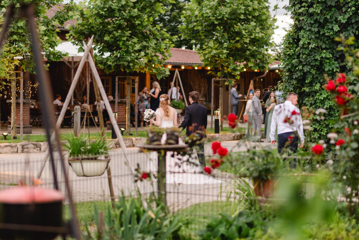 Braut und Bräutigam spazieren durch die Gartenanlage des Hochzeits-Schammenhofs in Langenau. Im Hintergrund sind Gäste zu sehen. Grünpflanzen und rote Blumen rahmen die Szene ein. Braut und Bräutigam spazieren durch die Gartenanlage des Hochzeits-Schammenhofs in Langenau. Im Hintergrund sind Gäste zu sehen. Grünpflanzen und rote Blumen rahmen die Szene ein.