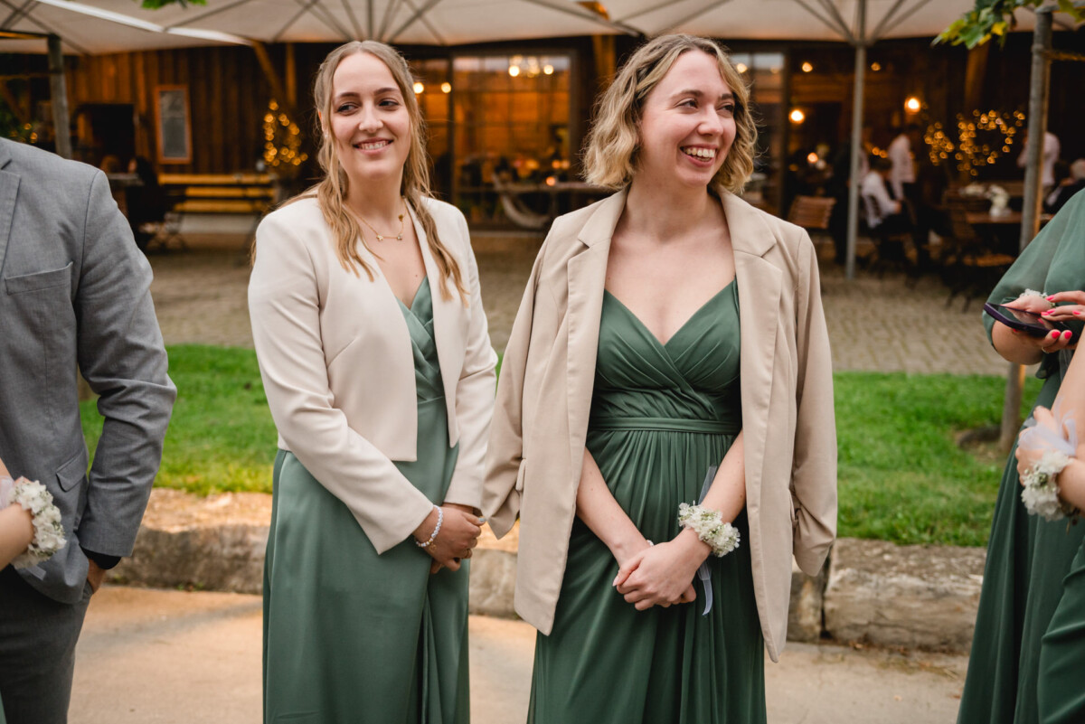 Zwei Frauen in grünen Kleidern und beigen Jacken stehen lächelnd draußen auf dem Hochzeitsplatz Schammenhof in Langenau bei Ulm. Im Hintergrund sind Menschen und Lichter zu sehen, die die fröhliche Atmosphäre verstärken. Zwei Frauen in grünen Kleidern und beigen Jacken stehen lächelnd draußen auf dem Hochzeitsplatz Schammenhof in Langenau bei Ulm. Im Hintergrund sind Menschen und Lichter zu sehen, die die fröhliche Atmosphäre verstärken.