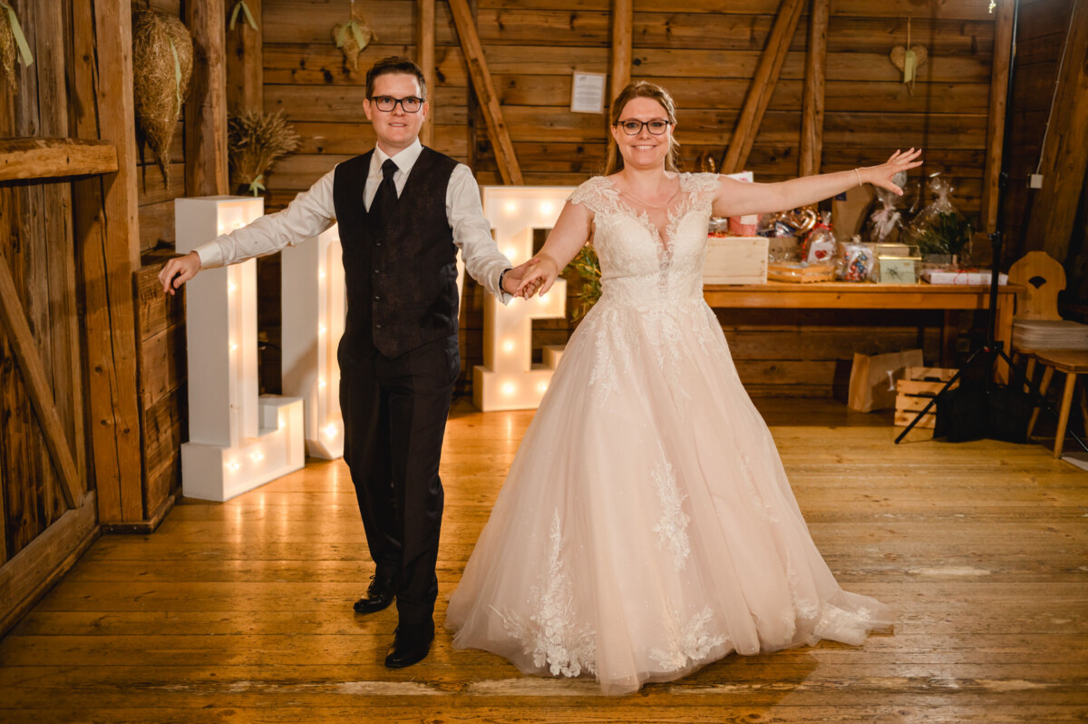 Braut und Bräutigam tanzen gemeinsam in einem Holzsaal auf dem Hochzeit Schammenhof bei Langenau. Im Hintergrund leuchten sanft Leuchtbuchstaben. Beide lächeln und strecken die Arme nach außen. Braut und Bräutigam tanzen gemeinsam in einem Holzsaal auf dem Hochzeit Schammenhof bei Langenau. Im Hintergrund leuchten sanft Leuchtbuchstaben. Beide lächeln und strecken die Arme nach außen.