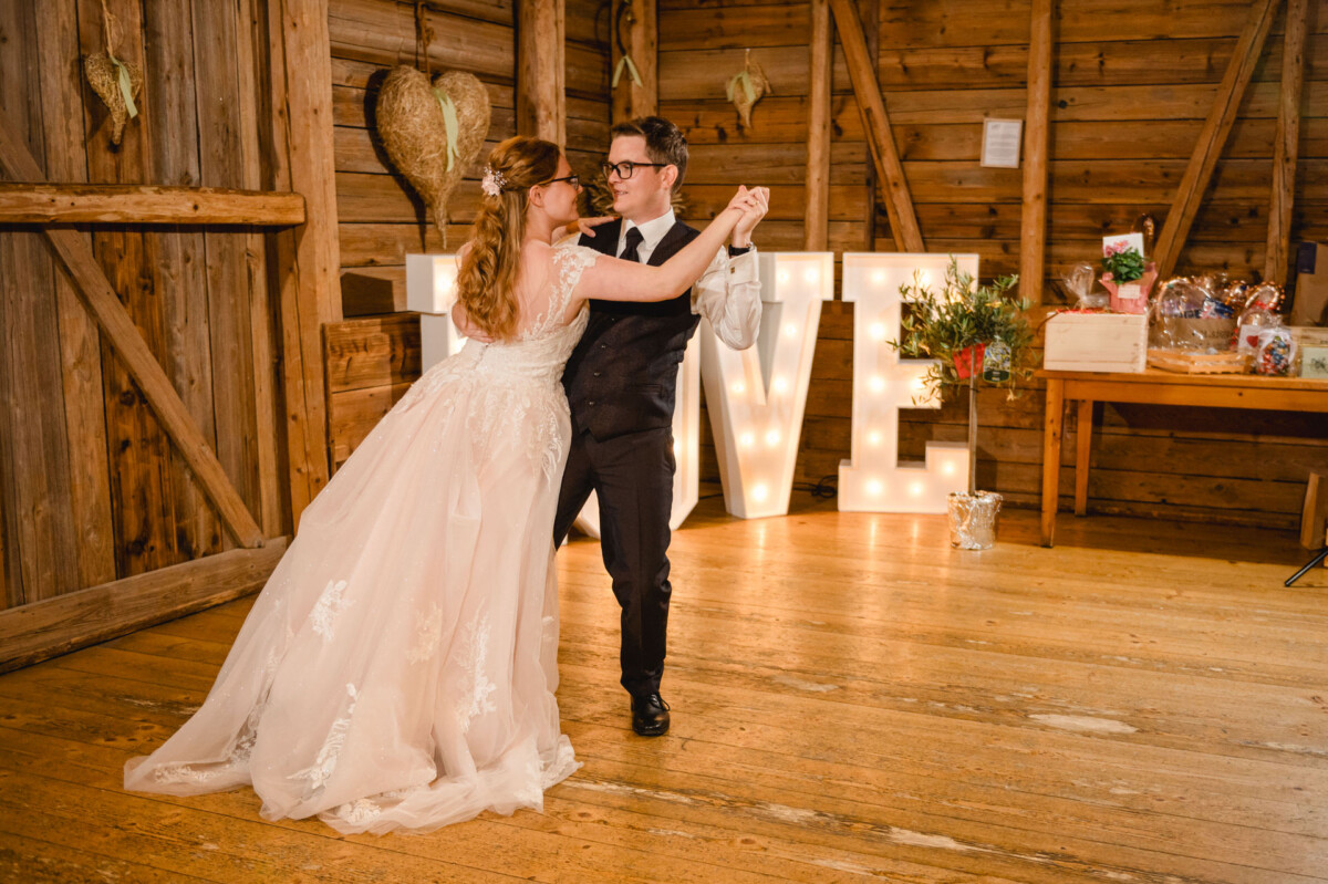 Ein Paar tanzt in einer Holzscheune auf dem Schammenhof, im Hintergrund sind große beleuchtete Buchstaben mit der Aufschrift „LOVE“ zu sehen und sorgen für eine zauberhafte Hochzeit in der Nähe von Langenau und Ulm. Ein Paar tanzt in einer Holzscheune auf dem Schammenhof, im Hintergrund sind große beleuchtete Buchstaben mit der Aufschrift „LOVE“ zu sehen und sorgen für eine zauberhafte Hochzeit in der Nähe von Langenau und Ulm.