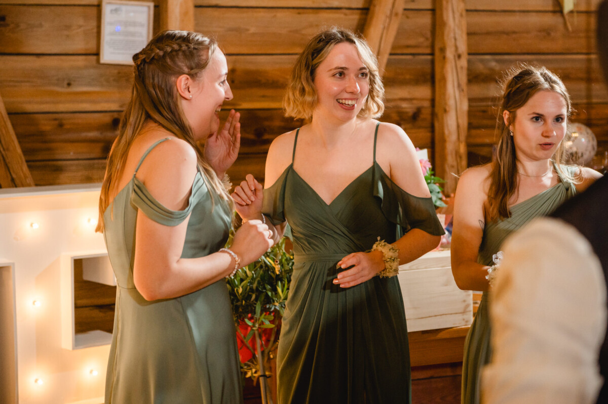 Drei Frauen in grünen Kleidern stehen im Hochzeitssaal des Schammenhofs in Langenau, lächeln und unterhalten sich. Ein großer Leuchtbuchstabe und Holzwände verleihen der lebhaften Szenerie nahe Ulm Charme. Drei Frauen in grünen Kleidern stehen im Hochzeitssaal des Schammenhofs in Langenau, lächeln und unterhalten sich. Ein großer Leuchtbuchstabe und Holzwände verleihen der lebhaften Szenerie nahe Ulm Charme.