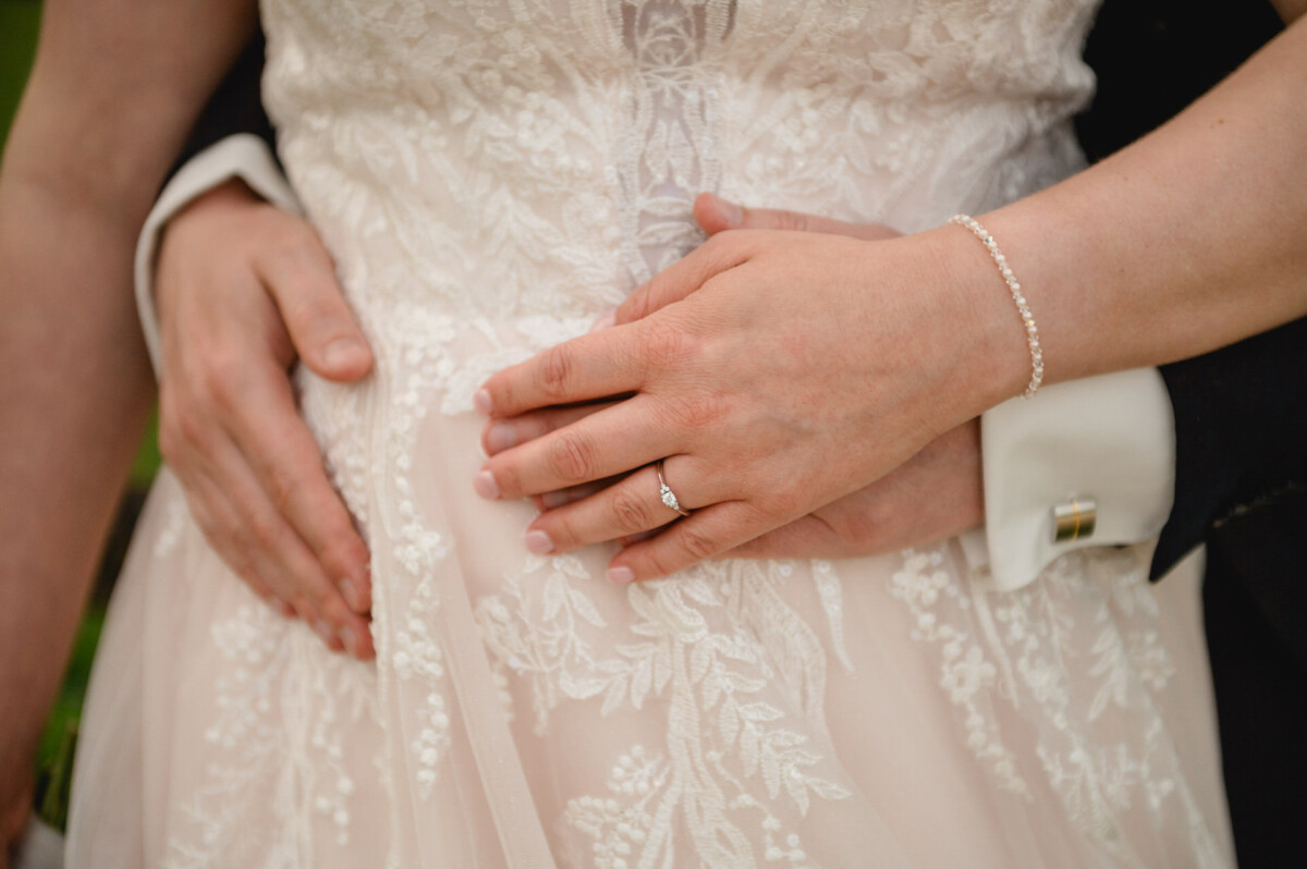 Nahaufnahme einer Person in einem Spitzenbrautkleid auf dem Hochzeit Schammenhof in Langenau bei Ulm. Ihre Hände sind um ihre Taille mit denen einer anderen Person verschränkt. Die Person trägt einen Ring und ein Armband. Nahaufnahme einer Person in einem Spitzenbrautkleid auf dem Hochzeit Schammenhof in Langenau bei Ulm. Ihre Hände sind um ihre Taille mit denen einer anderen Person verschränkt. Die Person trägt einen Ring und ein Armband.
